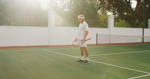 A Caucasian man wearing tennis whites spending time on a court, playing tennis on a sunny day, holding a tennis racket and celebrating, in slow motion.