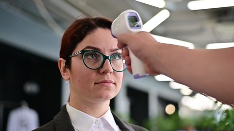 Temperature measurement by electronic infrared thermometer for office staff. Unhappy woman with a fever. A mandatory check at work with a thermometer gun is a non-contact forehead reading. Coronavirus
