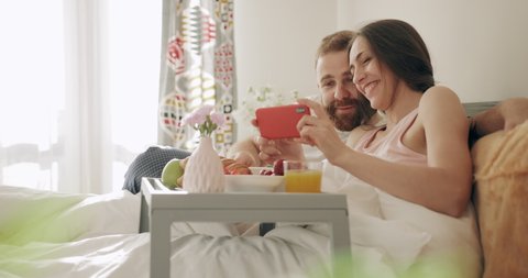 Cheerful family watching photos while sitting on bed with tray full of food. Young woman and man having breakfast in bed, laughing while looking and touching smartphone screen