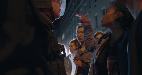 Angry female activist protesting against police violence. Young woman showing her frustration to a policeman while standing with group of protestors at night.
