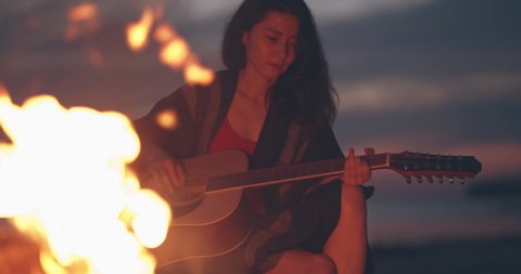 Solo activity and social distancing. Lonely girl sitting by bonfire on the beach near forest playing acoustic guitar. Spend free time in nature. Woman enjoying her loneliness. Summer vacation concept