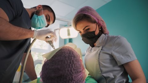 Beautiful Female Dentist in Protective Shield and Face Mask with the Help of an Male Assistant and Medical Equipment Works with Patient in Dental Office. Healthcare and Medicine Concept. Lower angle.