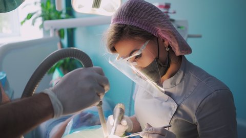Beautiful Female Dentist in a Protective Shield and Face Mask with the Help of an Male Assistant and Medical Equipment Works with a Patient in Dental Office. Healthcare and Medicine Concept