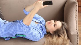 The girl poses on the phone camera, takes a selfie. A young woman lies on the sofa in her apartment and smiles. The blonde takes her own photos. Video to your smartphone.Close-up portrait