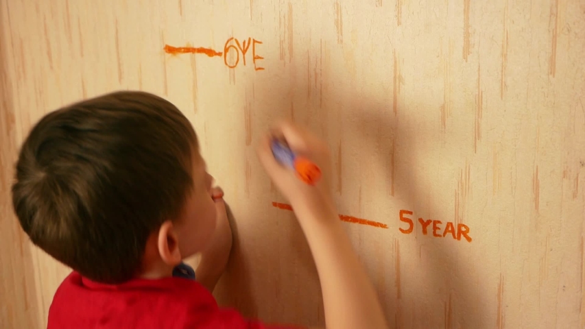 Concept on the theme of growing up children. Happy Caucasian boy writes at the growth mark on the wall his age is 6 years old, also on the wall is a mark with the inscription 5 years old. Birthday 6 y | Shutterstock HD Video #1054383203