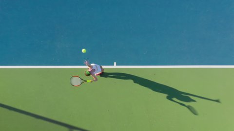 4K aerial top-down view of a young female sportsman during her practice. A close-up of a girl athlete serves the tennis ball. Young woman is hitting the ball with her tennis racket at sunset