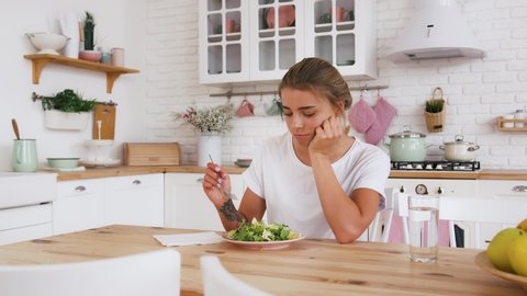 Lady sitting at table, feeling sad and bored with diet not wanting to eat salad. Modern kitchen interior. Healthy nutrition, eating disorder. Close up