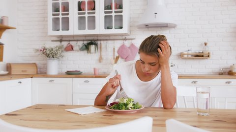 Woman sitting at table, looking sad and bored with diet not wanting to eat salad. Modern kitchen interior. Healthy nutrition, dieting, eating disorder
