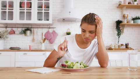 Female sitting at table, feeling unhappy with diet not wanting to eat salad. Modern kitchen interior. Healthy nutrition, eating disorder. Close up