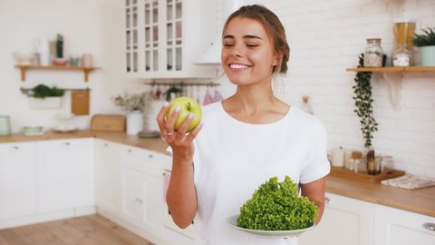 Woman is throwing up apple and eating it, holding green salad leaves. Modern kitchen interior. Healthy nutrition, eating disorder. Slow motion