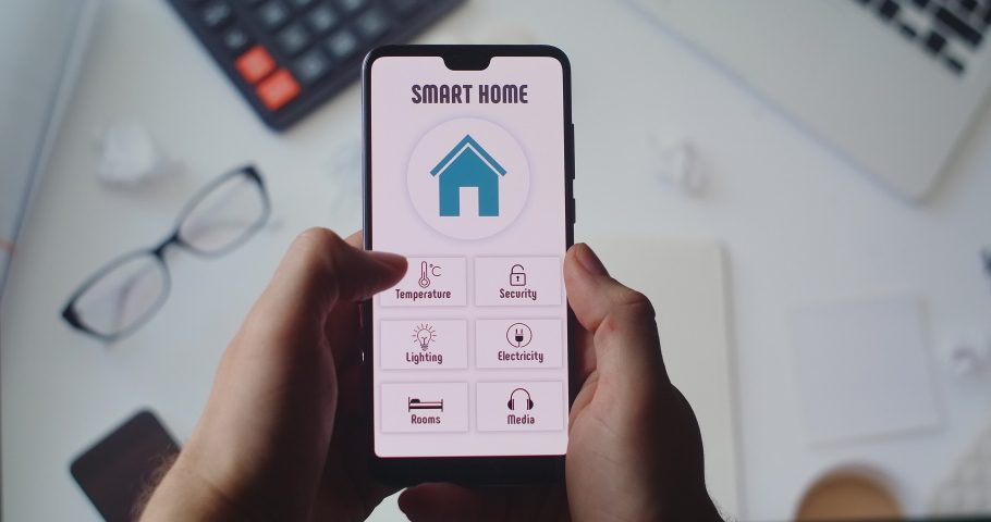 Manage smart home technologies from the office. On the background of the desktop, hands hold a smartphone with the smart home app and set the temperature and alarm in the house Royalty-Free Stock Footage #1054389248