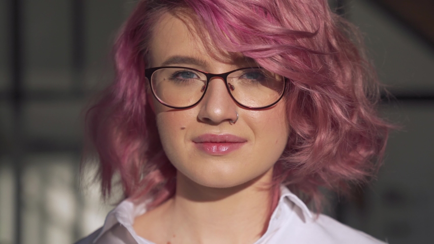Happy young adult hipster gen z teen girl smiling face with pink hair and nose piercing wearing glasses looking at camera posing indoors in modern sunny home office. Head shot close up portrait. | Shutterstock HD Video #1054390409