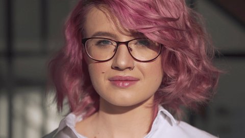 Happy young adult hipster gen z teen girl smiling face with pink hair and nose piercing wearing glasses looking at camera posing indoors in modern sunny home office. Head shot close up portrait.