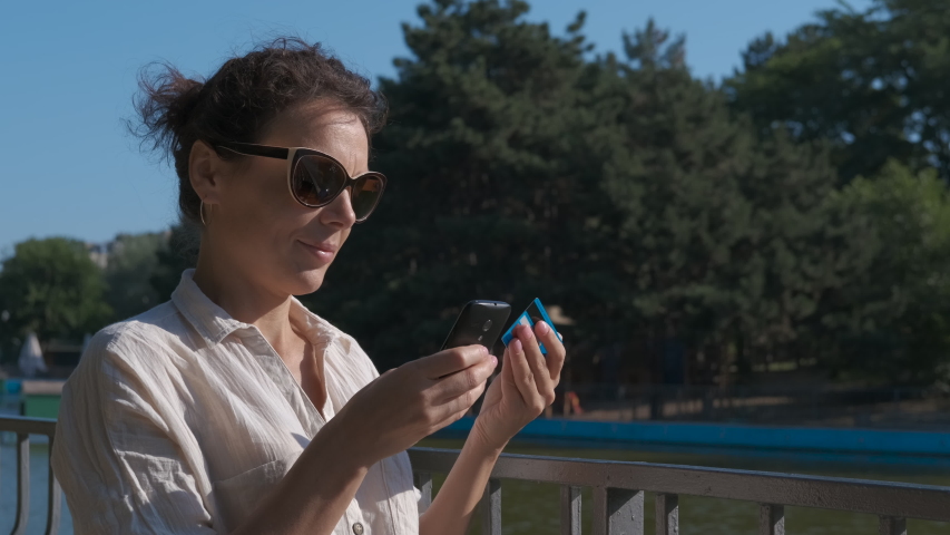 Put money on the phone. Happy woman in sunglasses pay something through phone on the city bridge.