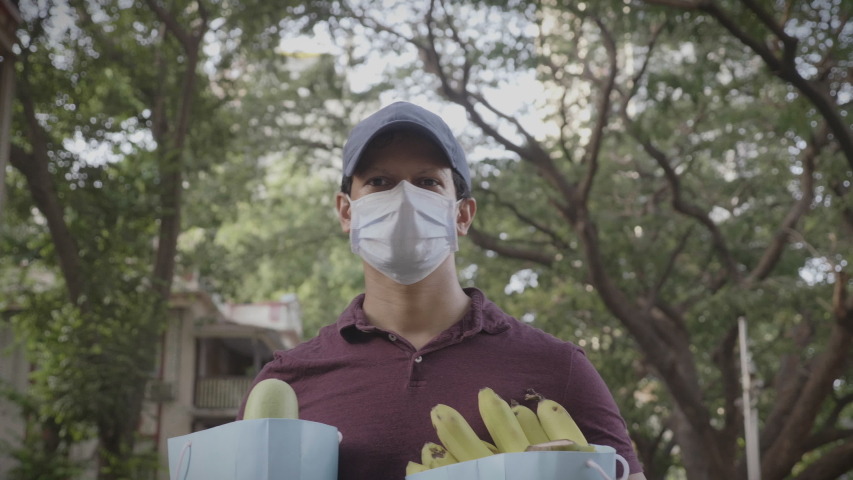 A movement shot of a delivery man boy wearing protective face mask carrying groceries fruits vegetables walking outdoors on the street road amid corona virus or COVID 19 epidemic or pandemic Royalty-Free Stock Footage #1054395056