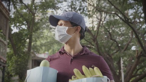 A movement shot of a delivery man boy wearing protective face mask carrying groceries fruits vegetables walking outdoors on the street road amid corona virus or COVID 19 epidemic or pandemic