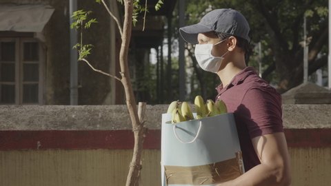 A delivery man boy wearing protective face mask and cap carrying groceries fruits vegetables walking outdoors on the street road amid corona virus or COVID 19 epidemic or pandemic