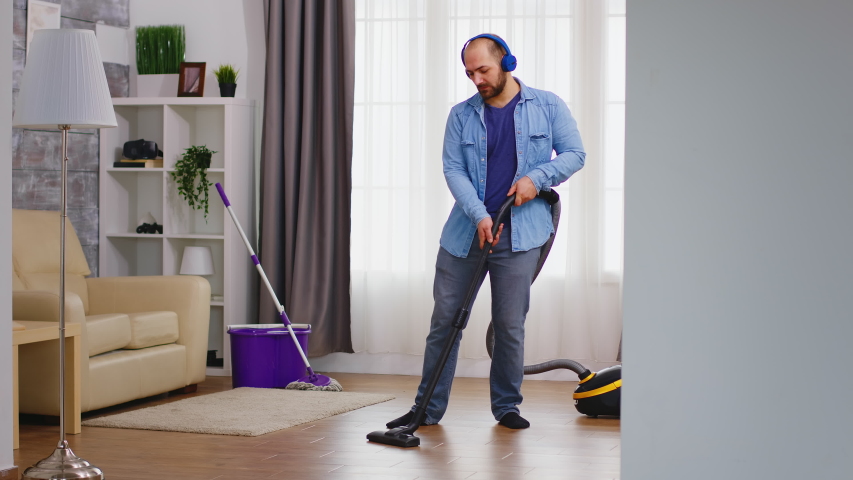 Funny young man listening music on headphone while cleaning the floor with vacuum cleaner. Happy dancing singing couple cleaning apartment, household and housekeeping, vaccum and mop, clean-up the hou | Shutterstock HD Video #1054398035