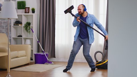 Funny young man listening music on headphone while cleaning the floor with vacuum cleaner. Happy dancing singing couple cleaning apartment, household and housekeeping, vaccum and mop, clean-up the hou