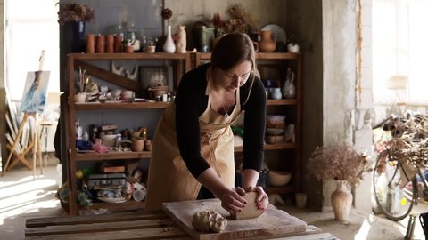 Front view of female potter wearing beige apron kneading softly clay piece on worktop, working with her hands. Pottery products on shelves behind, lens flares and sunlight from window. Slow motion