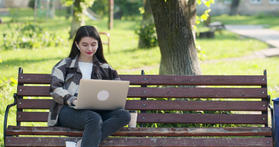 Caucasian woman in headset typing on the laptop computer on the bench in the park. Female student makes conference video call on laptop computer talks with web tutor, online teacher in webcam chat.