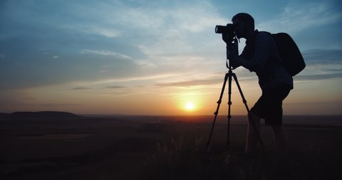 Silhouette of mature man in casual outfit with big backpack taking pictures of amazing sunset while standing on high hill. Photographer using professional tripod and digital camera outdoors.