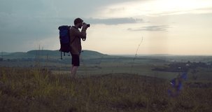 Full length portrait of travel photographer with backpack using digital camera for shooting nature while standing at green hill. Bearded man enjoying working process on fresh air.