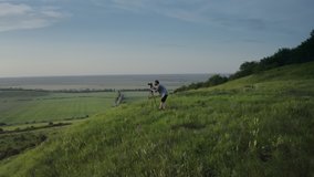 Male photographer standing on green hill with tripod and professional camera and making photos of beautiful landscape. Concept of adventure and nature.