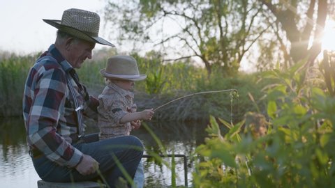 anglers, caring loving grandfather together with lovely happy grandson, fishing using a makeshift fishing rod sitting on pier by river on warm afternoon at sunset on weekend in countryside