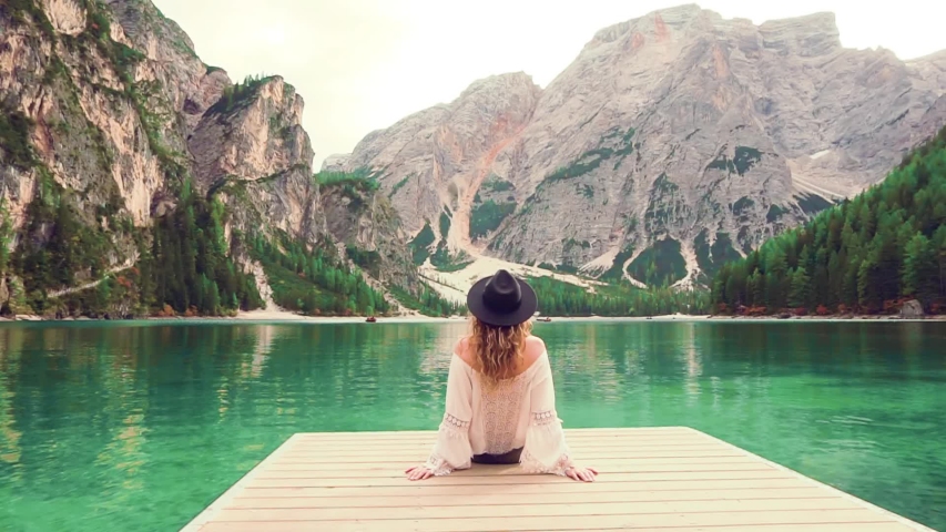 Mysterious woman tourist sits on wooden pier. Amazing view turquoise water high mountain lake Braies mountains. Casual boho style clothing cotton blouse shirt vintage black hat. turned away back view Royalty-Free Stock Footage #1054411829