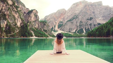 Mysterious woman tourist sits on wooden pier. Amazing view turquoise water high mountain lake Braies mountains. Casual boho style clothing cotton blouse shirt vintage black hat. turned away back view