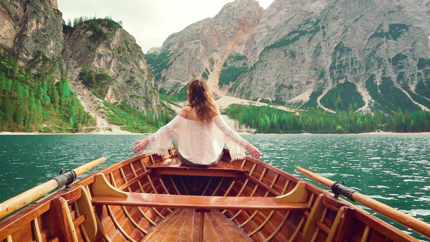 Young woman in vintage white boho shirt with blond hair loose turned away sitting on edge of wooden boat. Girl enjoy alpine azure lake clear water fabulous nature of mountain green forest Italian Alps Royalty-Free Stock Footage #1054411841