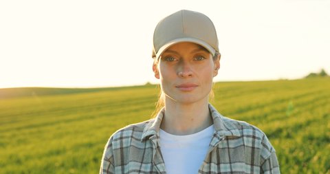 Portrait shot of attractive young Caucasian woman in hat standing in green field, smiling cheerfully to camera and giving thumb up. Female farmer with smile outdoors in summer. Camera zooming out.