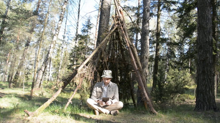 the guy sits in the forest in a makeshift shelter and looks at something on his tablet on a summer day hd