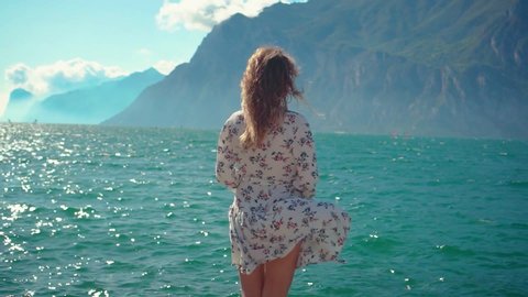 young woman with long hair white fluttering dress turned away stands on stone Riva del Garda old town waterfront enjoy sping mountains nature view lake Lago di Garda Trentino Alto Adige region Italy