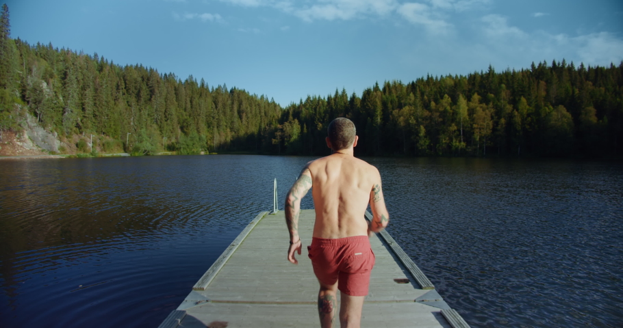Millennial young man with tattoos run on wooden dock or pier and jump into refreshing cool water of mountain forest lake. Concept staycation adventures and summer time activities in backyard  Royalty-Free Stock Footage #1054418219