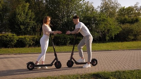 A young guy meets a beautiful girl in the Park, they ride electric scooters and talk. Slow motion. Teenagers have fun in the summer in the Park and ride electric scooters.