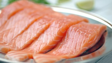 Close-up slow-motion of sliced fresh raw red salmon.
Raw salmon in a silver plate on the wooden table with lemon and herbs/greens ingredients on the side. Concept of healthy seafood. Salmon in 4K.
