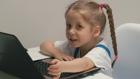 slow motion closeup portrait of little cute girl with pigtails sitting at table at home in living room behind laptop, on home online learning, kid looks into camera falsely smiling