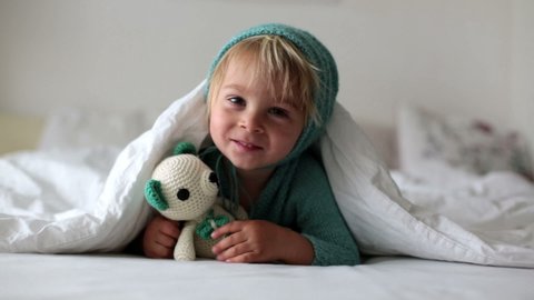 Cute toddler boy with handmade knitter toy, lying in bed, smiling happily