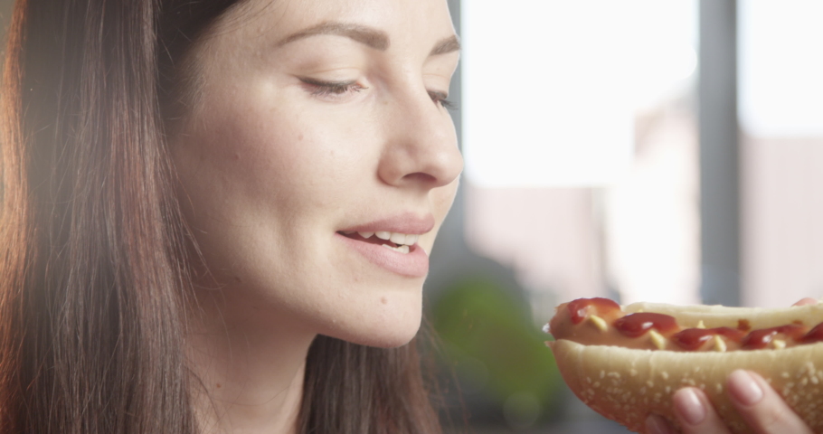 Hungry Woman Eating Hot Dog Enjoying the Taste Indoors with Sun Shining a Close up Shot on Red Camera Royalty-Free Stock Footage #1054421168