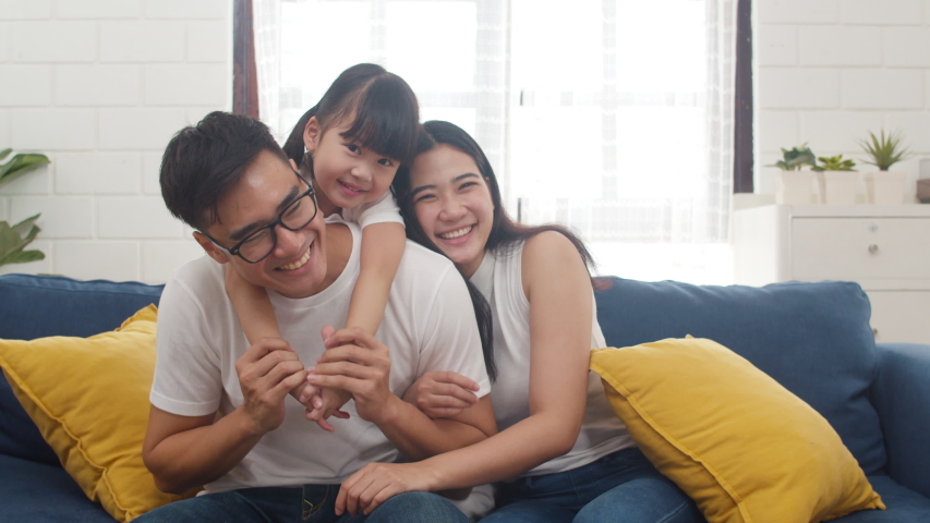Happy cheerful Asian family dad, mom and daughter having fun cuddling and video call on laptop on sofa at house. Self-isolation, stay at home, social distancing, quarantine for coronavirus prevention.