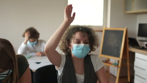 Curly masked schoolgirl pulls his hand up, ready to answer. Children in the class after quarantine coronavirus covid-19, back to school, post-quarantine life, new normality