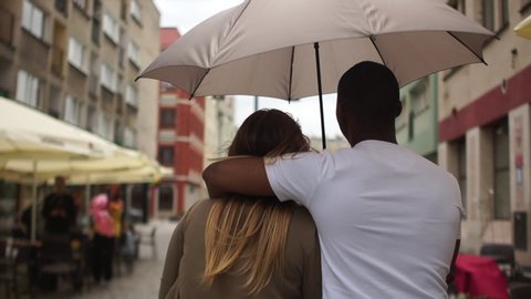 Black man holding an umbrella in his hands. Beautiful mixed race couple running along a city street while it is raining. Caucasian woman and black man, students couple in love. Back view