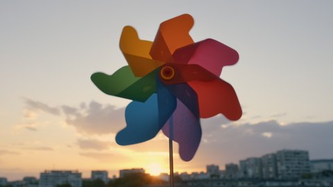Pinwheel rotating colored plastic with blowing wind against a blue sky, white clouds, silhouettes of city buildings on sunset slow motion. Symbol of freedom and life. Toy spinner. Bright disk of sun