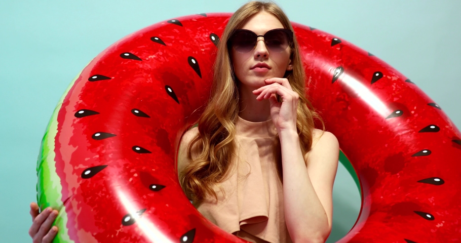 pensive beautiful girl in a summer hat and sunglasses, dressed in a swimsuit, stands with an inflatable ring on an blue background