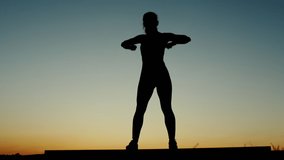 Young woman doing arm mobility exercises before starting jump stretches and squats silhouetted against a colorful sunset sky in a low angle view in a health and fitness concept