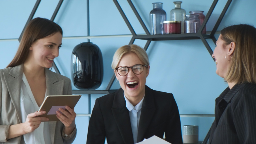 Three beautiful business women are standing in office and discussing project, idea, plan. Cheerful caucasian businesswomen laughing at workplace, different age generations in office Royalty-Free Stock Footage #1054428872