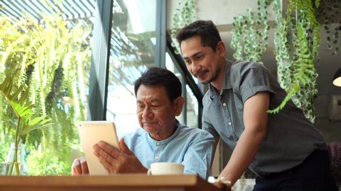 Portrait of a young man teaching his father how to use tablet computer. Son helping Dad to use the tablet until succeed and thumb up dad. Father and Son using a tablet together at cafe.