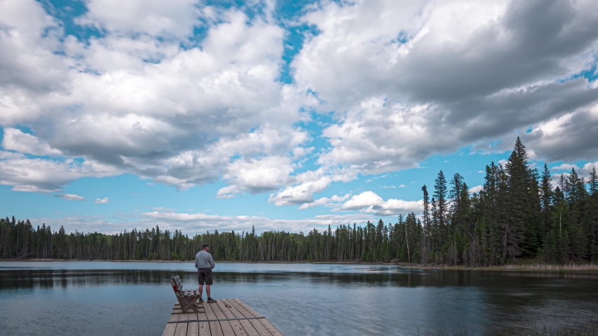 Time lapse of man fishing from a floating dock at a small lake.  A bright blue sky with blowing white clouds. 